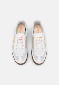 Bílé tenisky s texturovaným svrškem, zlatými třemi pruhy a oranžovým brandingem na béžových vložkách. Gumová podešev pro trakci.