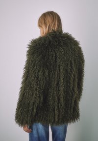 Oljčno zelena bunda iz umetnega krzna z voljno, teksturirano površino in ohlapnim krojem, ki ima visok ovratnik in široke rokave.