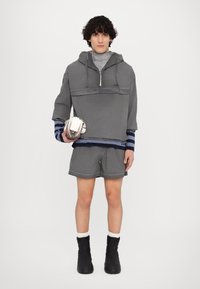 Hanorac gri cu glugă și fermoar scurt, pantaloni scurți asortați și botine negre până la gleznă. Prezintă dungi și un design texturat. Ține o geantă mică.