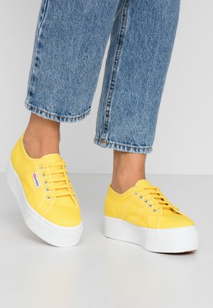 Osoba ubrana w krótkie niebieskie dżinsy i jasne żółte, na platformie, sznurowane trampki stojąca na jasnoszarej podłodze.