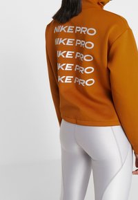 Pomarańczowa bluza Nike Pro z wysokim kołnierzem i powtarzającym się białym logo na plecach. W zestawie z błyszczącymi, jasnoszarymi legginsami.