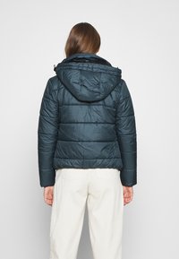 Mörkgrön puffjacka med huva, med ett quiltat mönster och elastiska ärmslut. Baksidan visar en kortad passform och en slät yta.