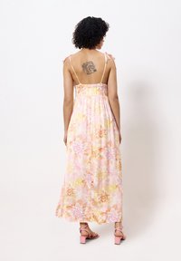 Robe maxi rose clair avec des motifs floraux en jaune, orange et violet. Présente des bretelles fines et ajustables ainsi qu'un dos smocké. Sandales à talons roses.