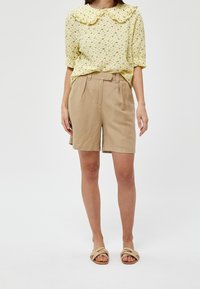 Blusa gialla a fantasia floreale con colletto arricciato, abbinata a pantaloni corti beige su misura. Materiale leggero, vestibilità casual, completata da sandali scivolati.