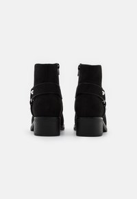 Botas de tobillo de ante negro con un bajo tacón cuadrado, cierre de cremallera y una correa decorativa con detalle de hardware plateado. Textura suave.