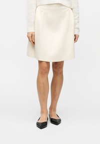 Γυναίκα με εκρού φούστα μέχρι τα γόνατα και πουλόβερ, όρθια, με μαύρες μυτερές μπαλαρίνες σε λευκό απλό φόντο.