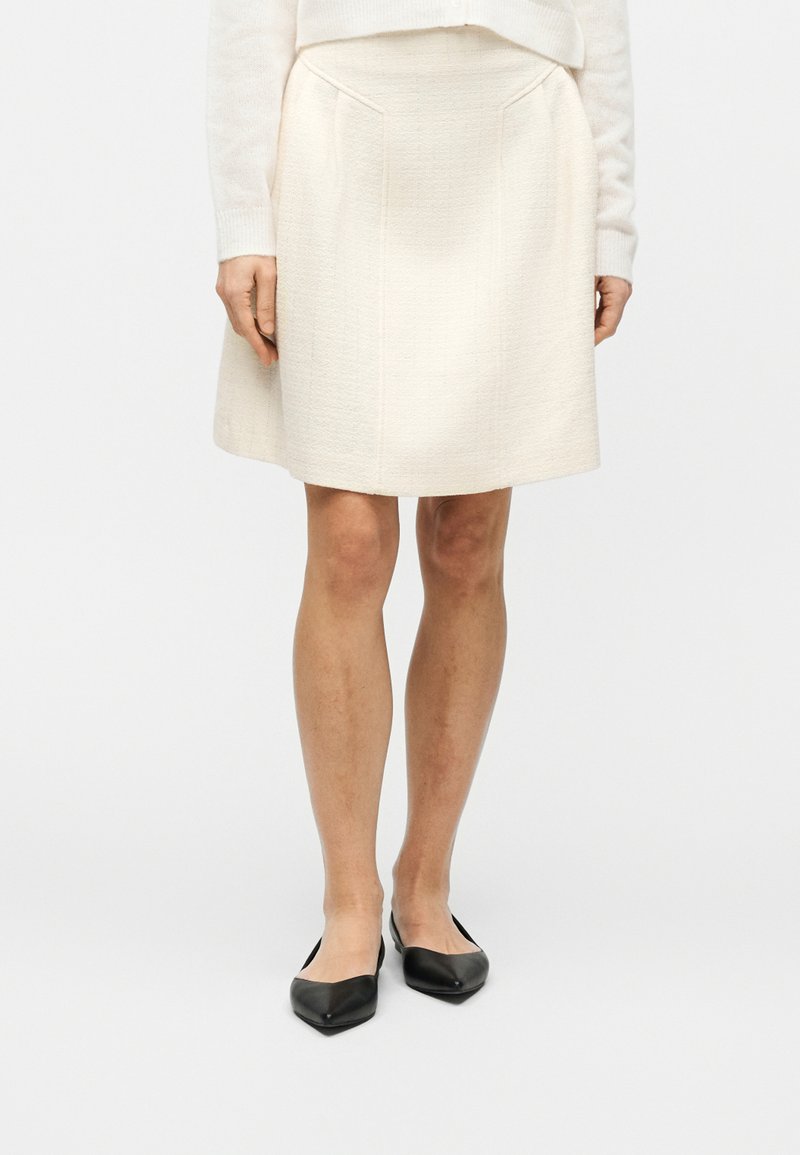 Γυναίκα με εκρού φούστα μέχρι τα γόνατα και πουλόβερ, όρθια, με μαύρες μυτερές μπαλαρίνες σε λευκό απλό φόντο.