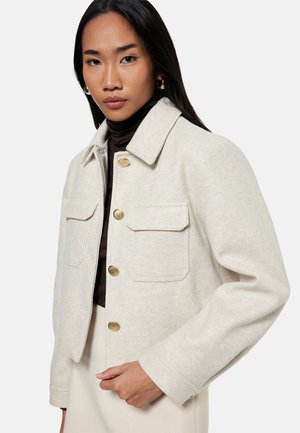 Frau trägt eine beige Wolljacke mit goldenen Knöpfen über einem dunklen Rollkragenpullover, steht vor einem schlichten hellen Hintergrund.