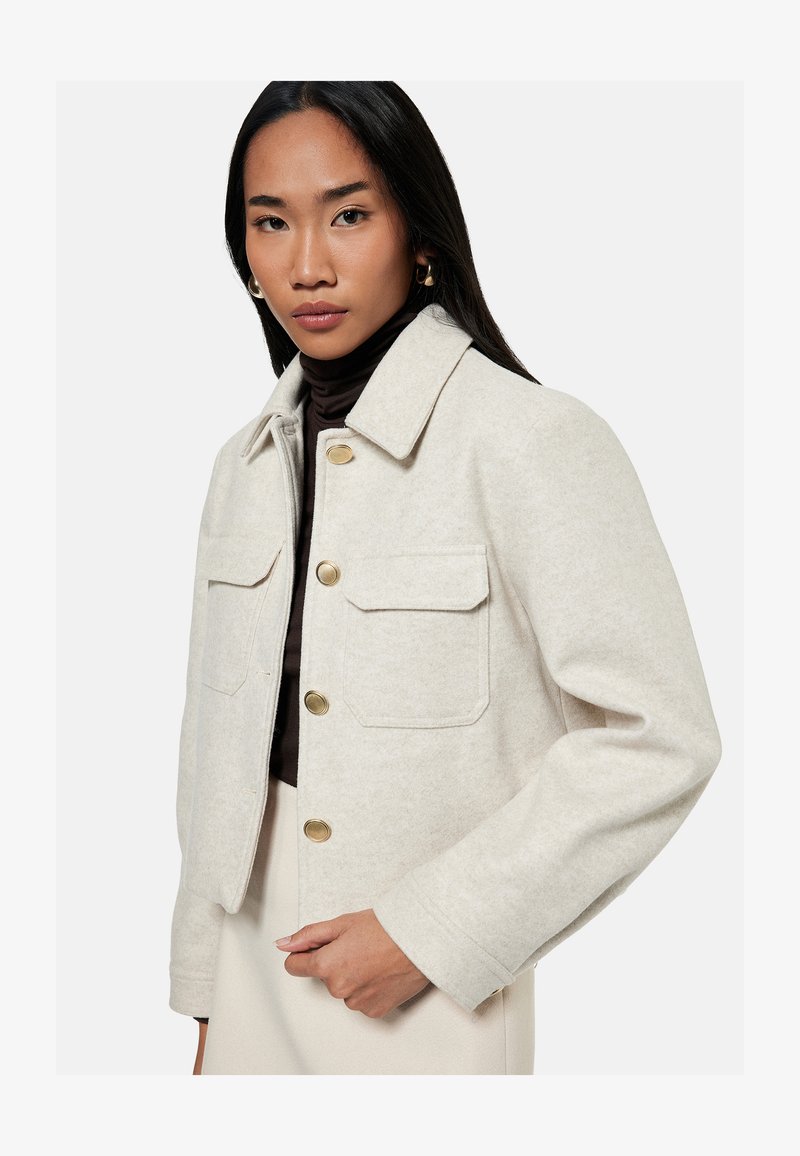 Vrouw die een beige wollen jas met gouden knopen draagt over een donkere coltrui, staand tegen een effen lichte achtergrond.
