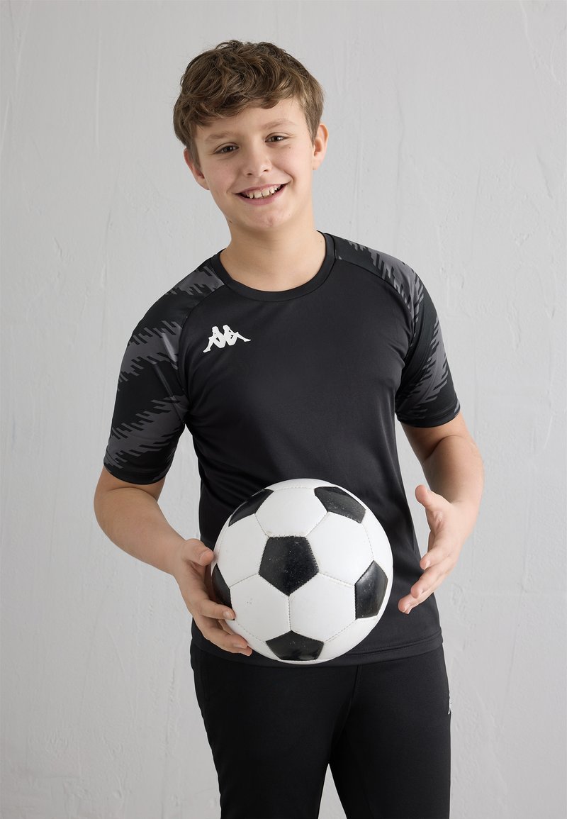 Usmívající se chlapec v černém sportovním oblečení držící černo-bílý fotbalový míč na jednoduchém bílém texturovaném pozadí.