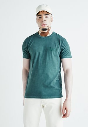 Camiseta de algodón verde con mangas cortas y cuello redondo, con un logotipo "Lee" en relieve en el pecho. Combinada con pantalones de color claro.