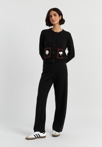 Una donna si erge con sicurezza in un cardigan nero con tasche decorative e pantaloni abbinati, indossando sneaker bianche con strisce nere.