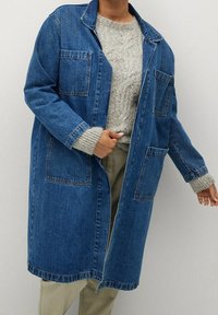 Person som bär en lång blå denimjacka över en grå kabelstickad tröja och beiga byxor, stående mot en enkel bakgrund.