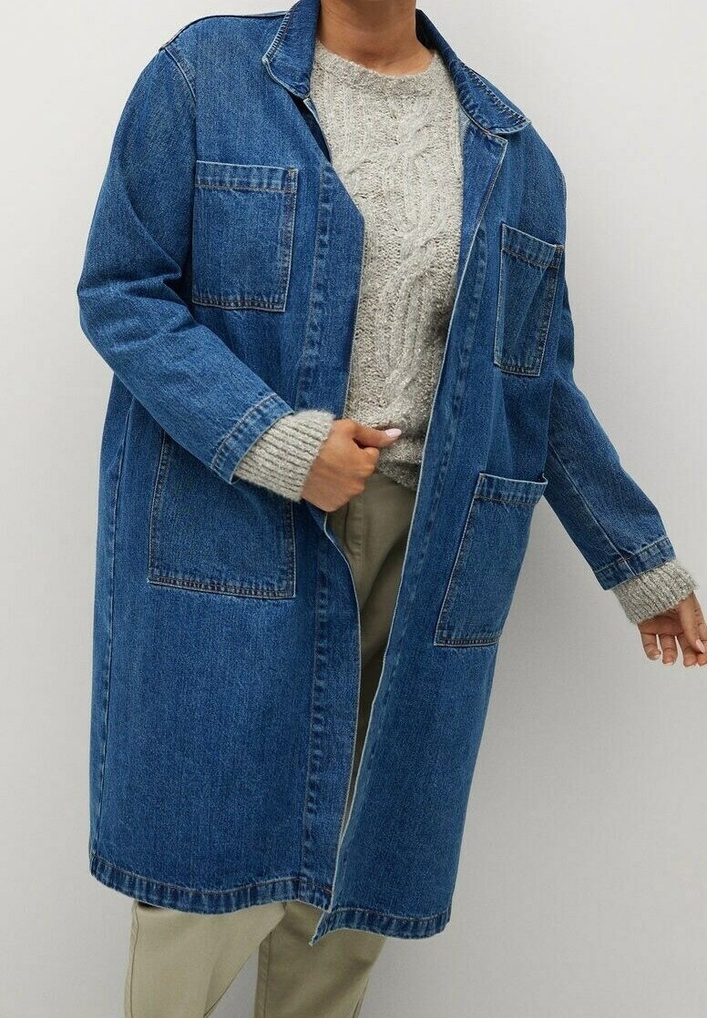 Person som bär en lång blå denimjacka över en grå kabelstickad tröja och beiga byxor, stående mot en enkel bakgrund.