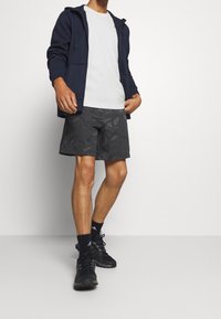 Man med marinblå zip-up hoodie, vit t-shirt, svarta mönstrade shorts, svarta Adidas-strumpor och svarta träningsskor som står mot en vit bakgrund.