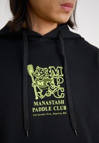 Black hooded sweatshirt featuring a yellow graphic of a bear with a paddle and text "MANASTASH PADDLE CLUB" and "710 Harbor Ave., Seattle, WA."