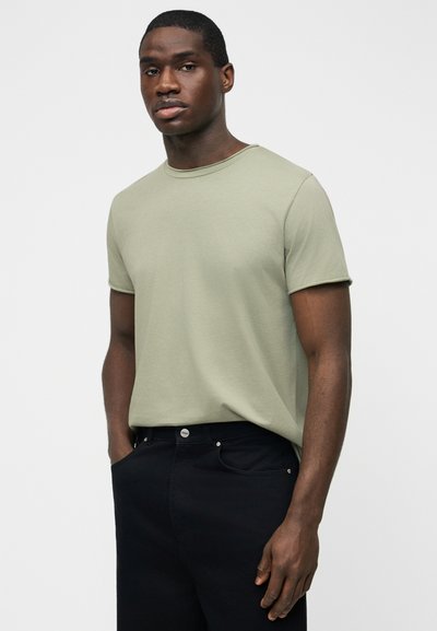 Άνδρας με κοντά μαλλιά που φοράει ανοιχτό πράσινο κοντομάνικο μπλουζάκι και μαύρο παντελόνι, στέκεται με τα χέρια στις τσέπες μπροστά σε απλό φόντο.