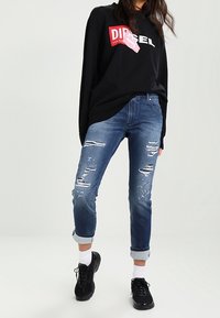 Sudadera negra de manga larga con el logo blanco de "DIESEL", vaqueros azules desgastados con roturas, y zapatillas negras con suelas blancas.
