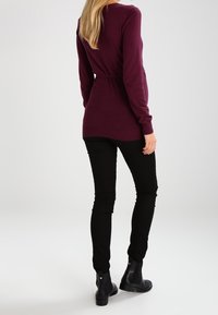 Długi burgundowy sweter z paskiem w talii, z długimi rękawami i zaokrąglonym dołem. Połączony z czarnymi spodniami slim-fit i botkami za kostkę.