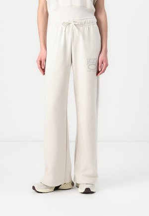 Personne portant un pantalon de jogging ample couleur blanc cassé avec taille élastique et logo Nike sur la cuisse gauche, assorti à des baskets assorties.