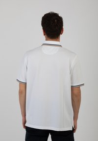 Camicetta bianca a polo con maniche corte, caratterizzata da un colletto a righe e un tessuto texturizzato. La vista posteriore mostra un'apertura circolare sul collo.