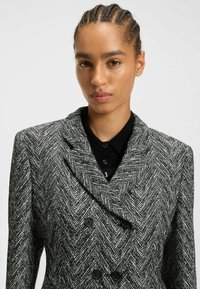 Dobbeltradet blazer i et sort og hvidt herringbone-mønster, med skrå krave, struktureret stof og sorte knapper.