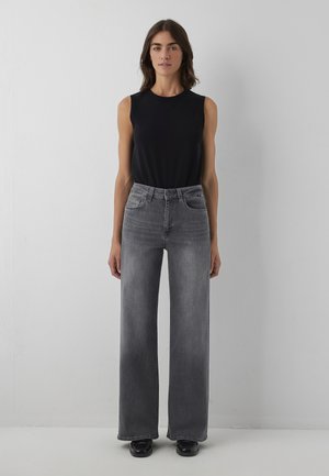 Femme portant un haut noir sans manches, un jean gris taille haute à jambes larges et des chaussures noires, debout devant un mur gris clair uni.