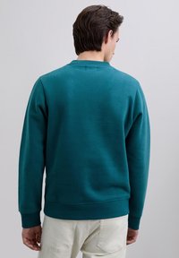 Hombre con sudadera de cuello redondo color verde azulado y pantalones blanco hueso, de pie de espaldas contra un fondo liso gris claro.