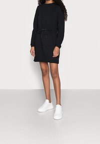 Svart långärmad klänning med dragsko i midjan; tillverkad av mjukt material. Kombinerad med vita sneakers mot en neutral bakgrund.