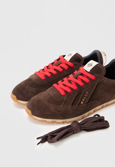 Sapatilhas de camurça castanha com atacadores vermelhos, sola de borracha natural e branding na língua. Inclui um par adicional de atacadores castanhos.