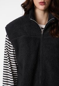 Crni prsluk od frotira s visokim ovratnikom, patentnim zatvaračem sprijeda i teksturiranim materijalom, slojevit preko prugaste dugih rukava.