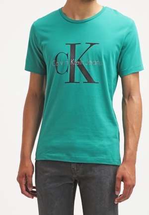 Homme portant un t-shirt Calvin Klein Jeans bleu sarcelle avec un grand logo "cK" noir et un jean gris, debout devant un fond blanc uni.