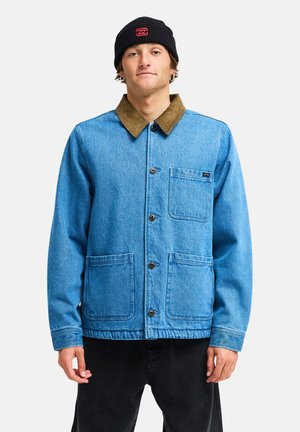 Jeune homme portant une veste en jean bleue avec un col marron, un bonnet noir, un pantalon noir et de petites boucles d'oreilles créoles, debout devant un fond uni.