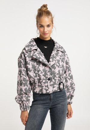 Junge Frau mit blondem Haar zu einem Dutt gebunden, trägt Creolen, eine rosa-schwarz gemusterte kurze Jacke und dunkle, hoch taillierte Jeans.