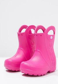 Bottes de pluie en caoutchouc roses avec des semelles texturées, des orteils ronds et une hauteur de cheville élevée. Équipées de poignées latérales pour un enfilage facile.