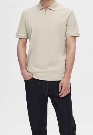 Mand iført beige polo med kort ærme og lynlås, krave og mørkeblå jeans, stående med den ene hånd i lommen mod hvid baggrund.