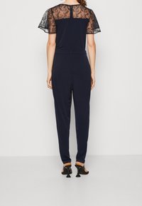 Combinaison bleu marine avec une superposition de dentelle sur les épaules, des manches courtes et une taille froncée. Elle présente des jambes fuselées et est portée avec des chaussures à talons.