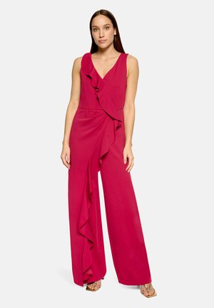 Frau steht und trägt einen ärmellosen, tiefrosa Jumpsuit mit weitem Bein und großem Rüschen-Detail vorne, kombiniert mit goldenen offenen Pumps.