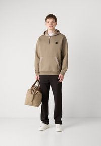 Sudadera caqui con media cremallera, bolsillo tipo canguro y detalles en beige, combinada con pantalones negros y una bolsa marrón claro, llevada con zapatillas blancas.