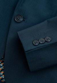 Marineblauwe kostuumstof met een gestructureerd patroon; heeft een drie-knopen manchet van glanzend zwart plastic en een geometrische printvoering in oranje en blauw.