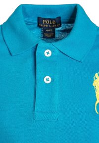 Turkos polo skjorta gjord av texturerat tyg. Har en krage, tre-knapp placket och en gul broderad logo på vänster sida.