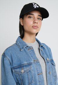 Jasnoniebieska kurtka dżinsowa z metalowymi guzikami i dwiema kieszeniami na piersi, zestawiona z szarym t-shirtem i czapką w kolorze czarnym z białym logo.