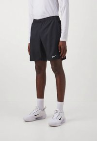 Άτομο που φορά μαύρες αθλητικές σορτς Nike, λευκό μακρυμάνικο μπλούζα, λευκά αθλητικά παπούτσια Nike και λευκές κάλτσες, στέκοντας σε λευκό πάτωμα.