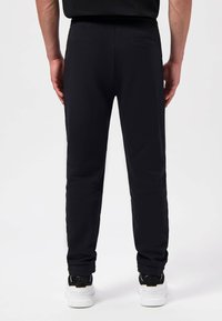 Zwarte joggers met een slim fit, elastische tailleband en boorden. Glad materiaal met een achterzak en zijnaaddetails. Draagt met witte sneakers.