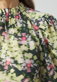 Femme portant un chemisier fleuri avec un col froncé et des manches bouffantes aux épaules, en vert, rose, noir et blanc.