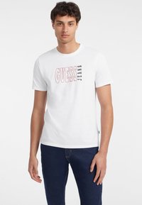 Balta medvilnė T-palaidinė su raudona ir juoda "GUESS JEANS" grafika. Dizainas paprastas, apvaliu iškirpimu ir trumpomis rankovėmis.