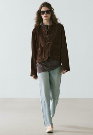 Donna che indossa una giacca oversize marrone scuro, top grigio, jeans azzurri, ballerine beige e grandi occhiali da sole neri mentre cammina in avanti.