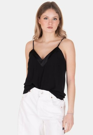 Giovane donna con capelli castano chiaro indossa un top nero senza maniche con scollo a V e pantaloni bianchi, in piedi davanti a uno sfondo semplice.