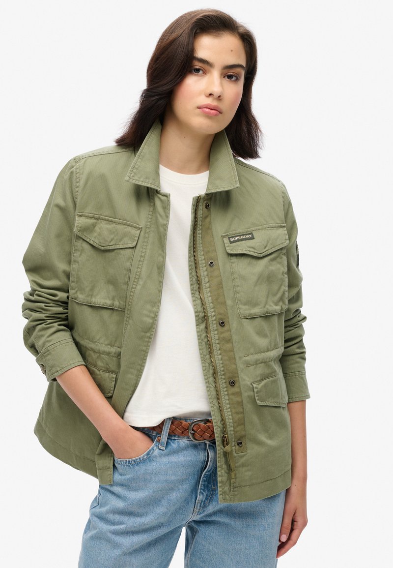 Olijfgroene, knoopsluiting jack met vier voorzakken, opgerolde mouwen en een subtiele stoftextuur. Draagt over een wit t-shirt en lichtblauwe jeans.