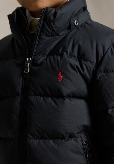 Niño con una chaqueta acolchada negra con un logo de caballo bordado en rojo en el pecho y un cuello parcialmente cerrado con cremallera.